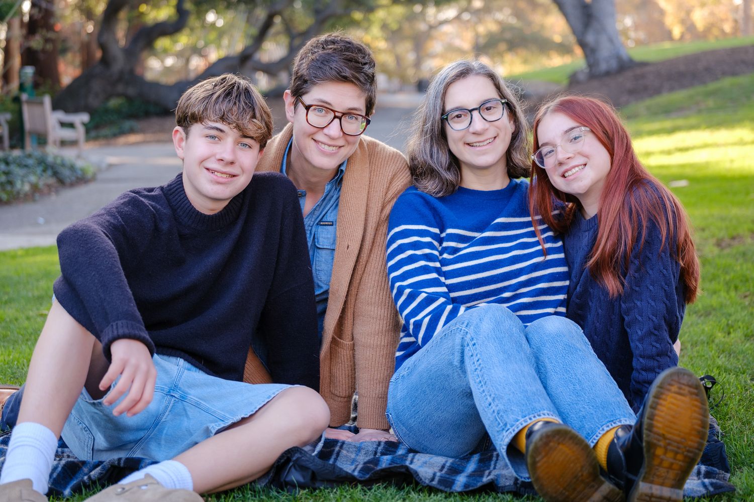 Four smiling teenagers sitting together on grass in a sunny park, posing for a casual outdoor group photo.