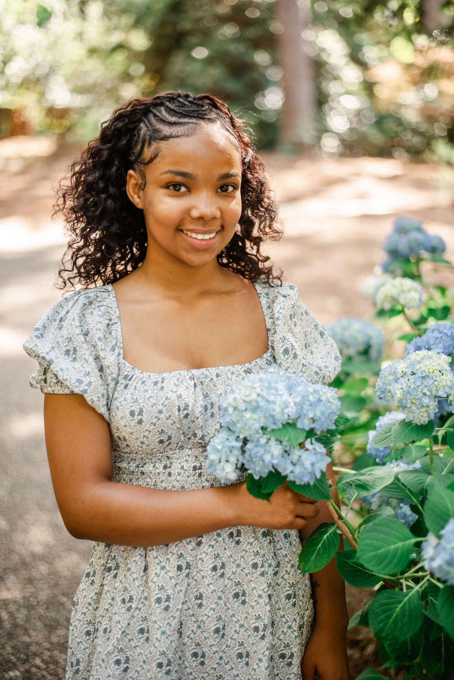 Young woman in floral dress smiling while holding blue hydrangeas in a sunlit garden.