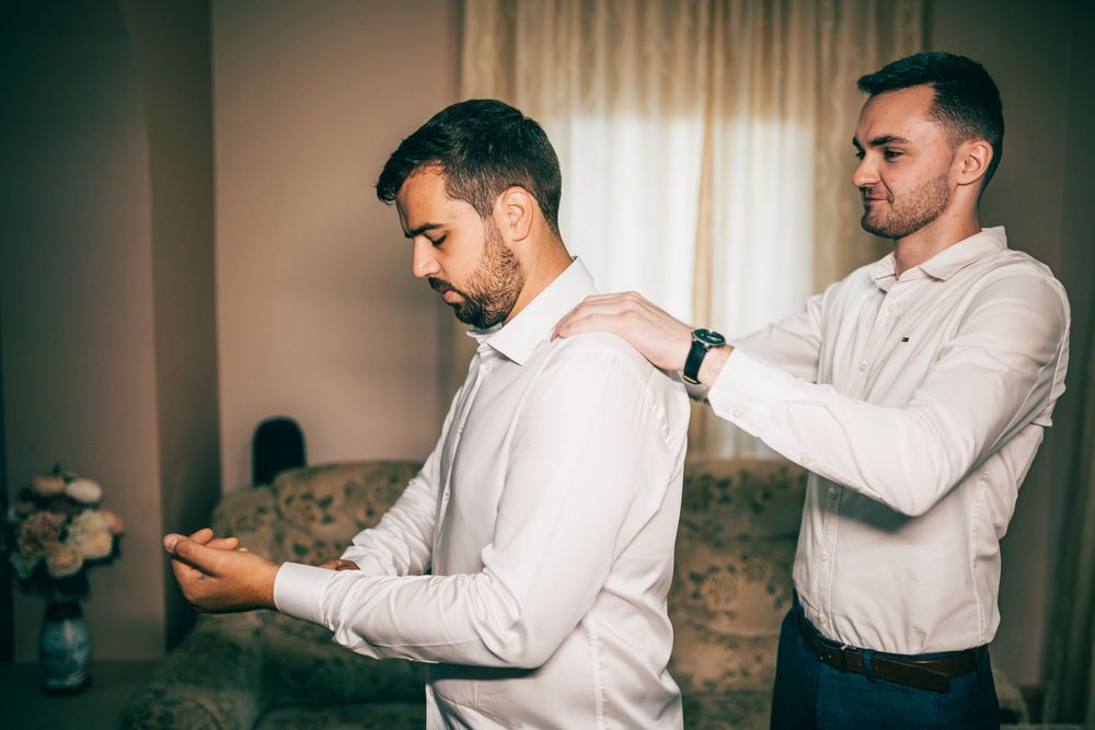 Groomsman helps groom button his white dress shirt while getting ready for wedding ceremony.