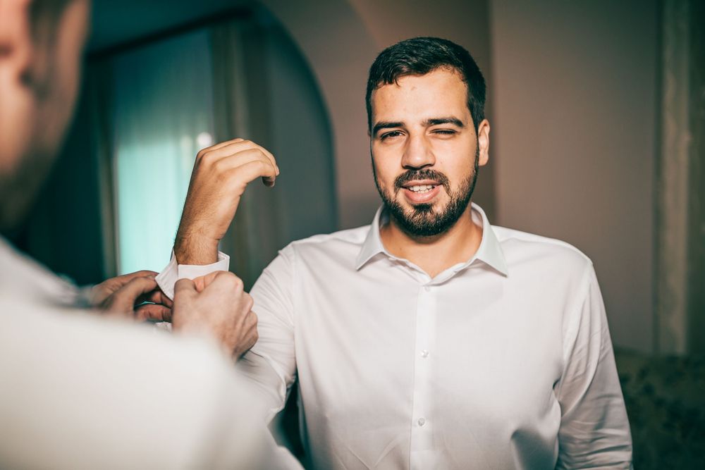 Groom in white shirt getting cufflinks fastened while smiling in mirror reflection before wedding.