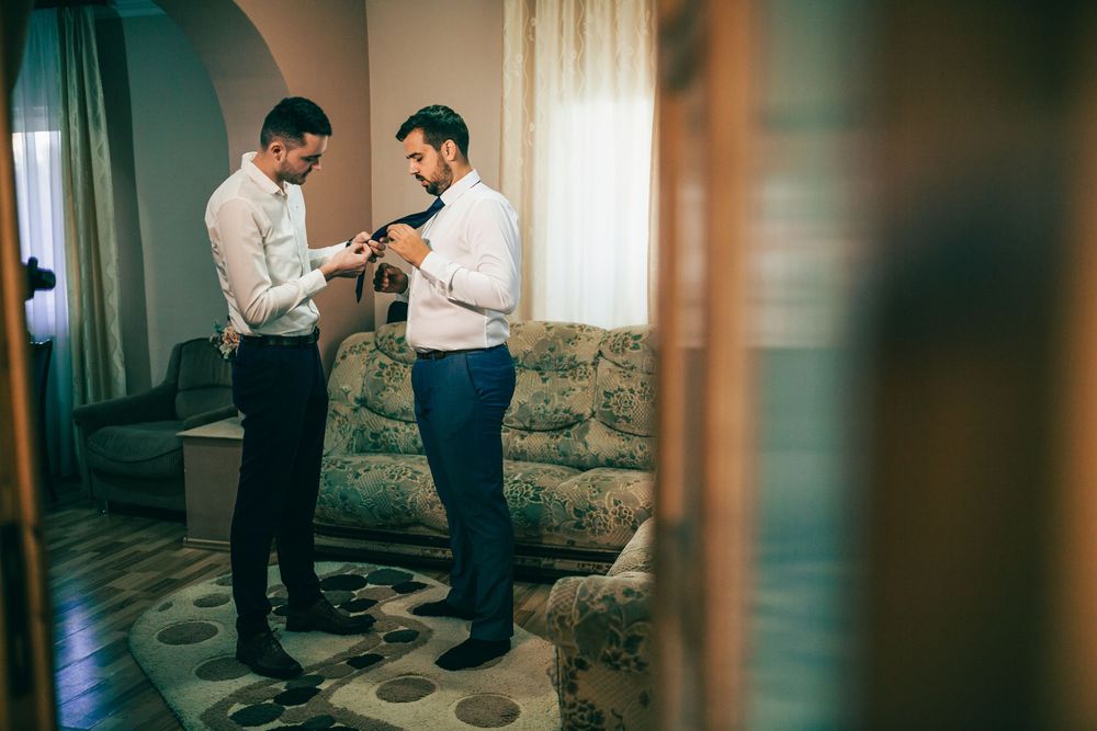 Two men in formal attire help each other prepare with neckties before a wedding ceremony.