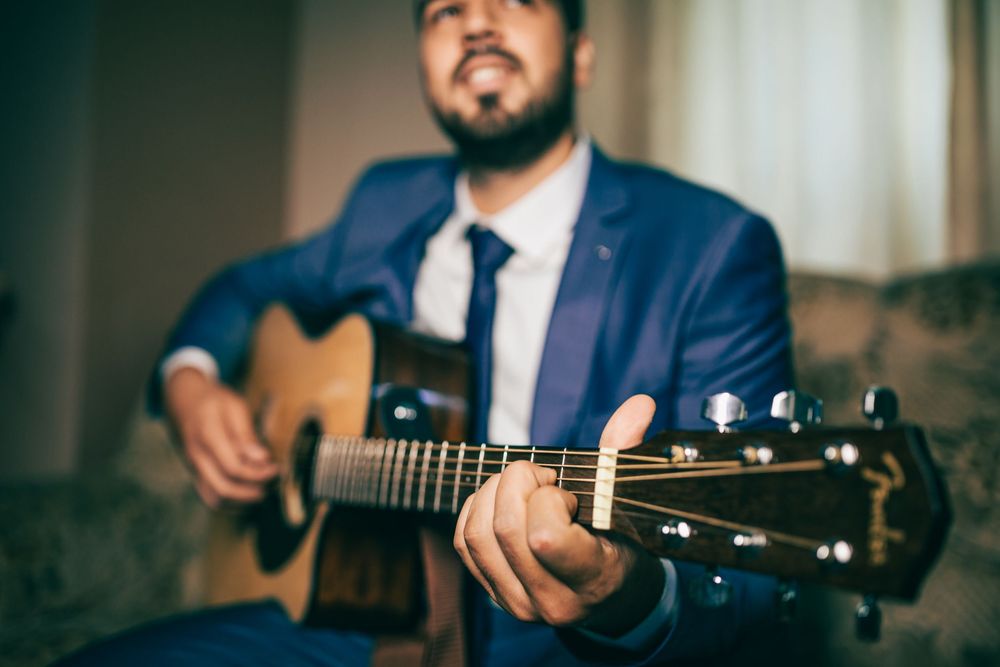 Bearded man in blue suit playing acoustic guitar while seated indoors.