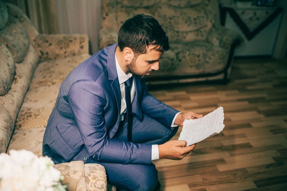 Groom in blue suit reads letter while sitting on sofa surrounded by wedding flowers.