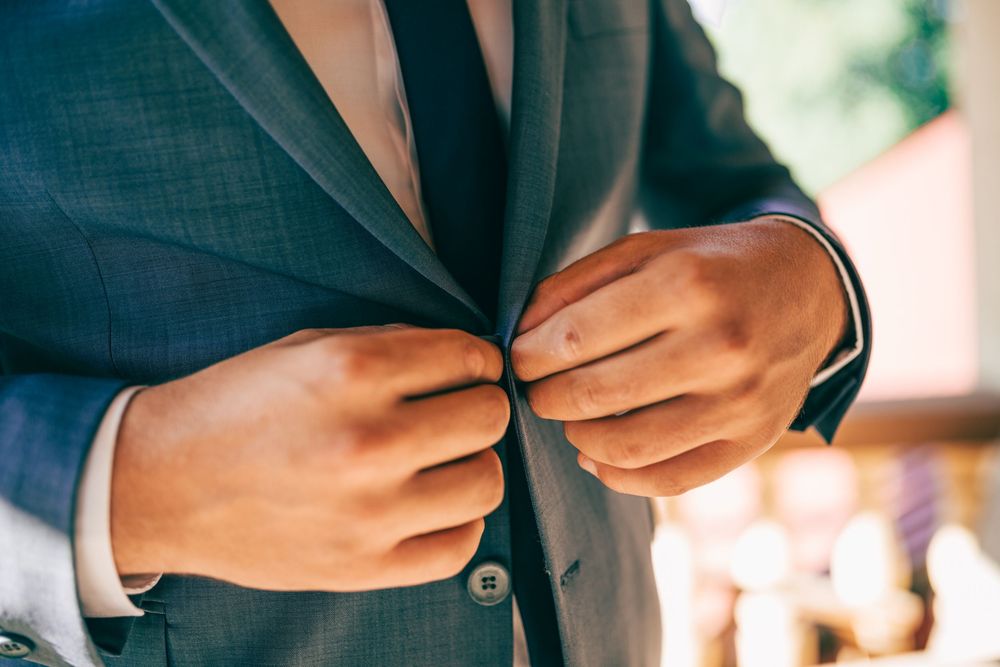 Groom in a blue suit buttoning his jacket during wedding day preparations in outdoor natural light.