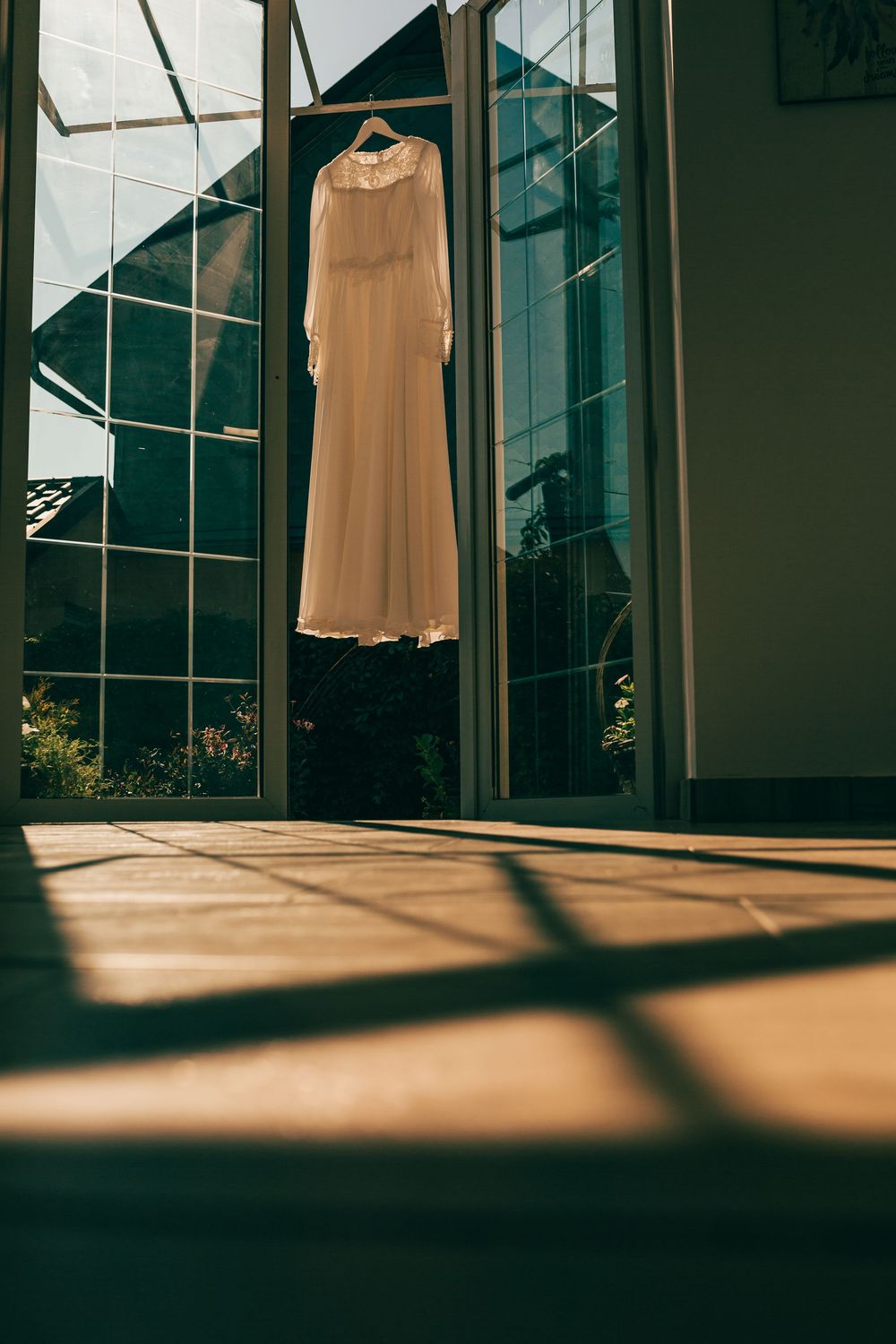 Elegant white wedding dress hanging on a door frame bathed in warm sunlight with glass windows in background.