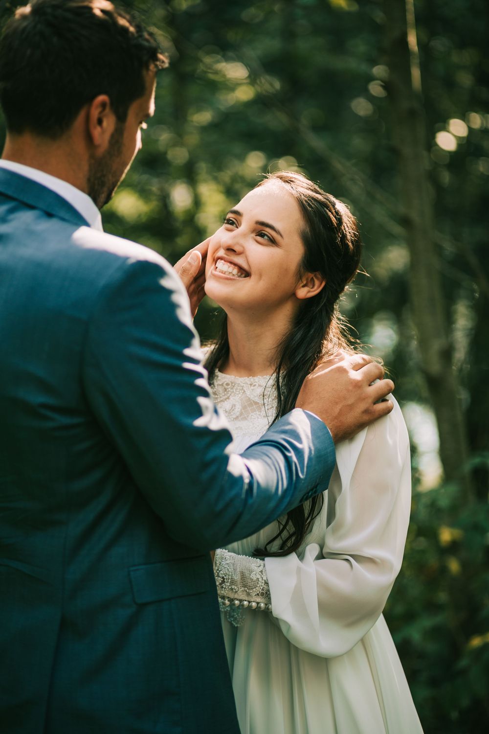 Smiling bride in white dress gazing at groom in blue suit outdoors in green forest setting.