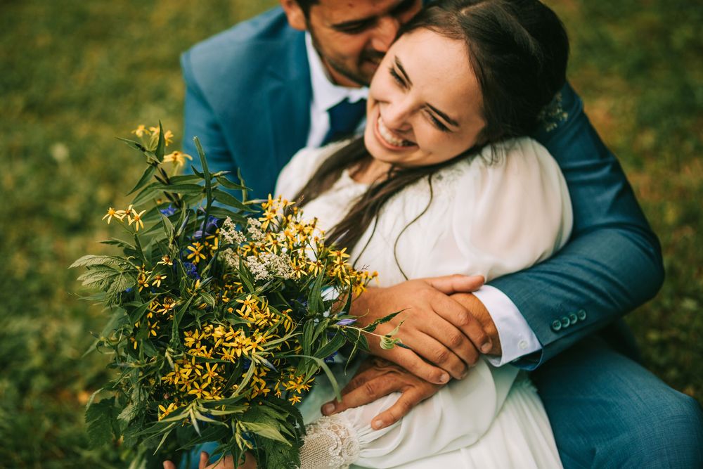 Bride and groom embracing outdoors holding yellow wildflower bouquet on green grass at wedding celebration.