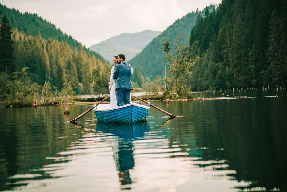 Newlyweds embracing in a blue rowboat on a serene mountain lake surrounded by lush green forests.