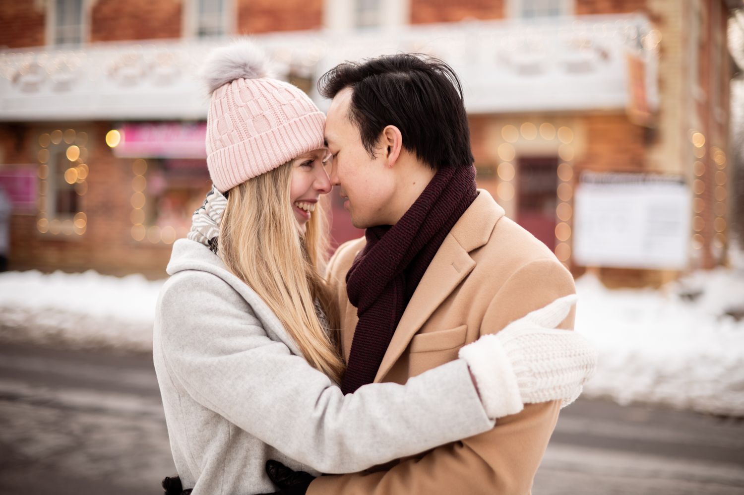A romantic couple embraces and touches foreheads outdoors in winter, wearing cozy scarves and hats in a snowy urban setting.