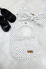 White polka dot baby bib and matching bow headband set displayed with black baby shoes on marble surface.