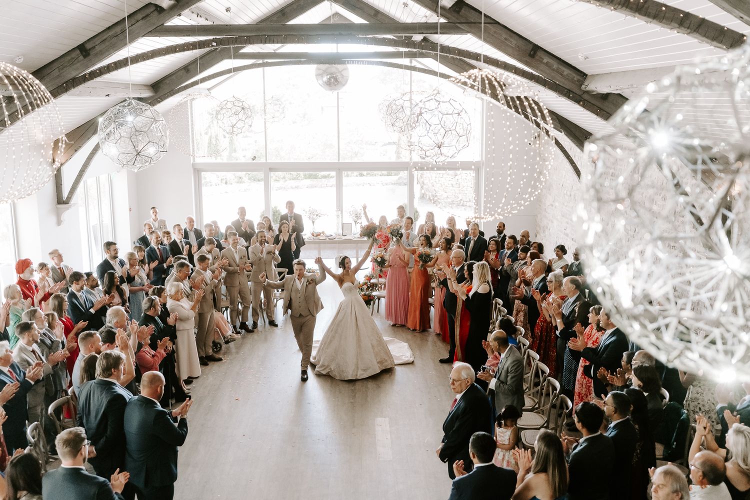 Bride and groom share first dance surrounded by cheering guests in elegant white barn wedding venue with sparkling chandeliers.