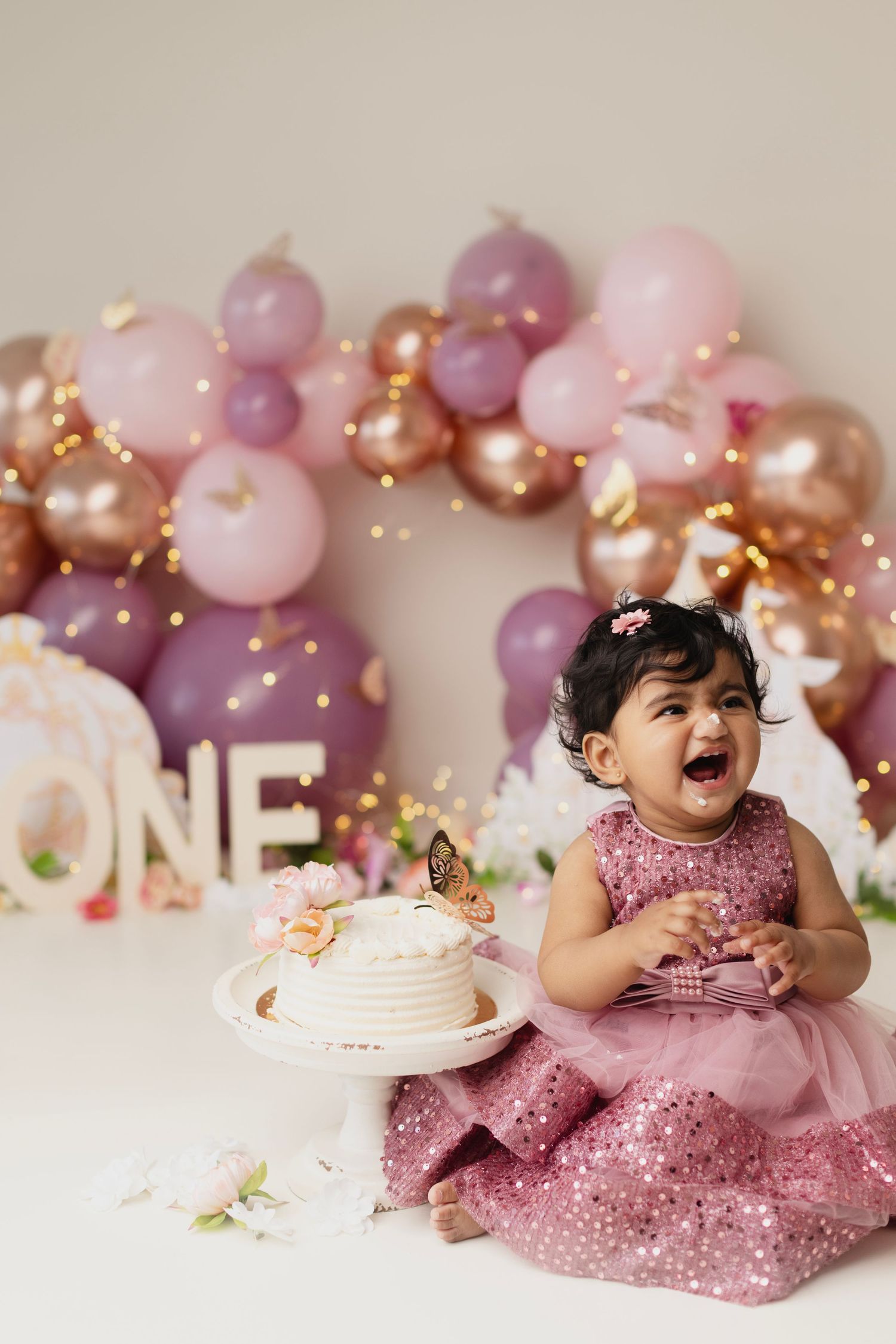 Baby girl in pink dress laughs at first birthday cake smash with balloon arch and ONE sign backdrop.
