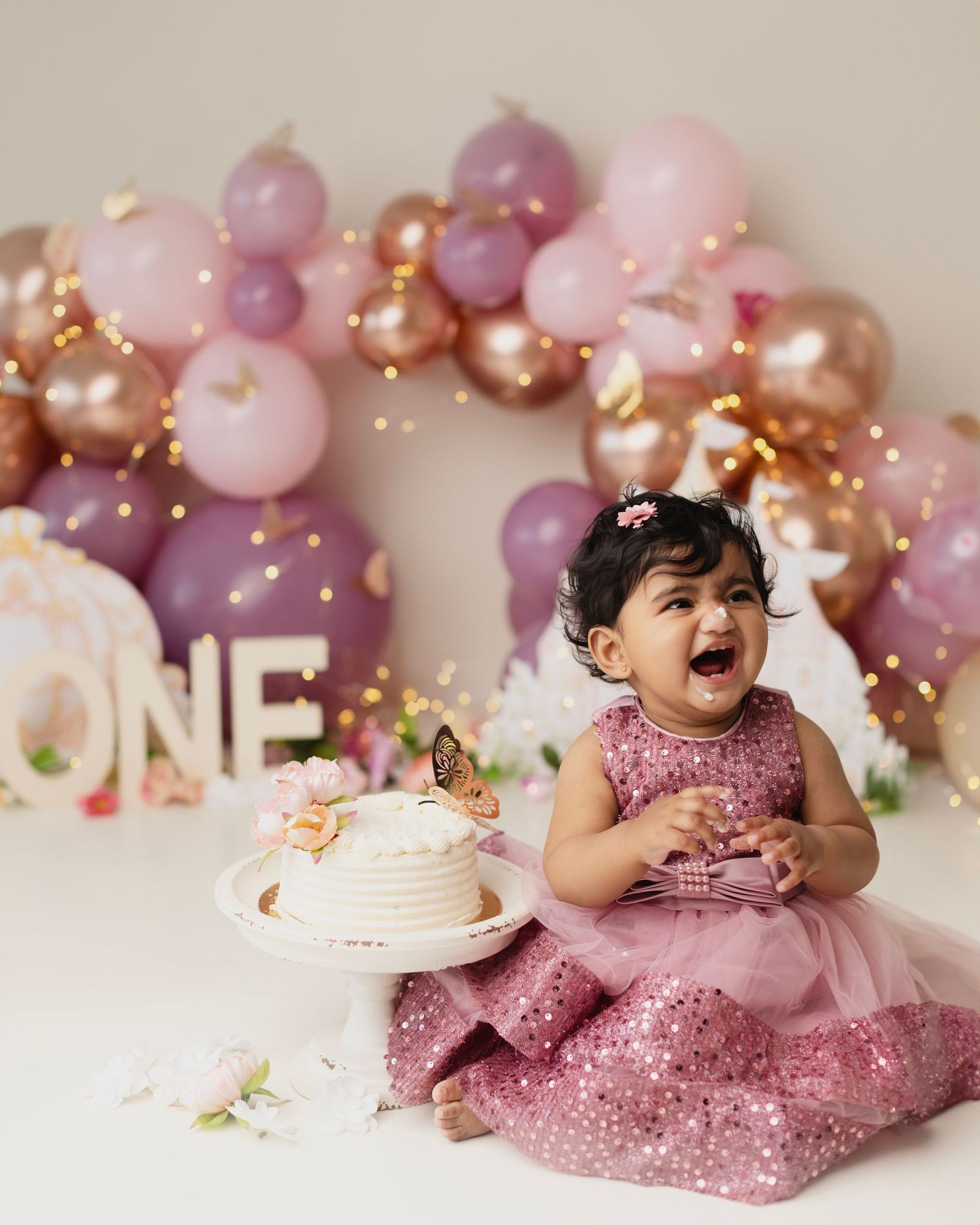 Baby girl in pink dress laughs at first birthday cake smash with balloon garland backdrop spelling ONE.