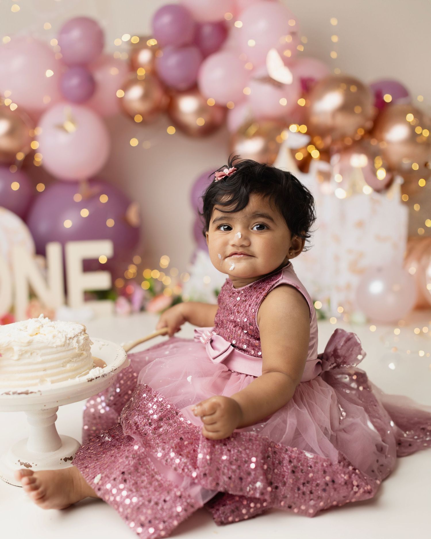 A baby girl in a pink sequin dress sits beside a white cake at her first birthday party with balloon decorations.