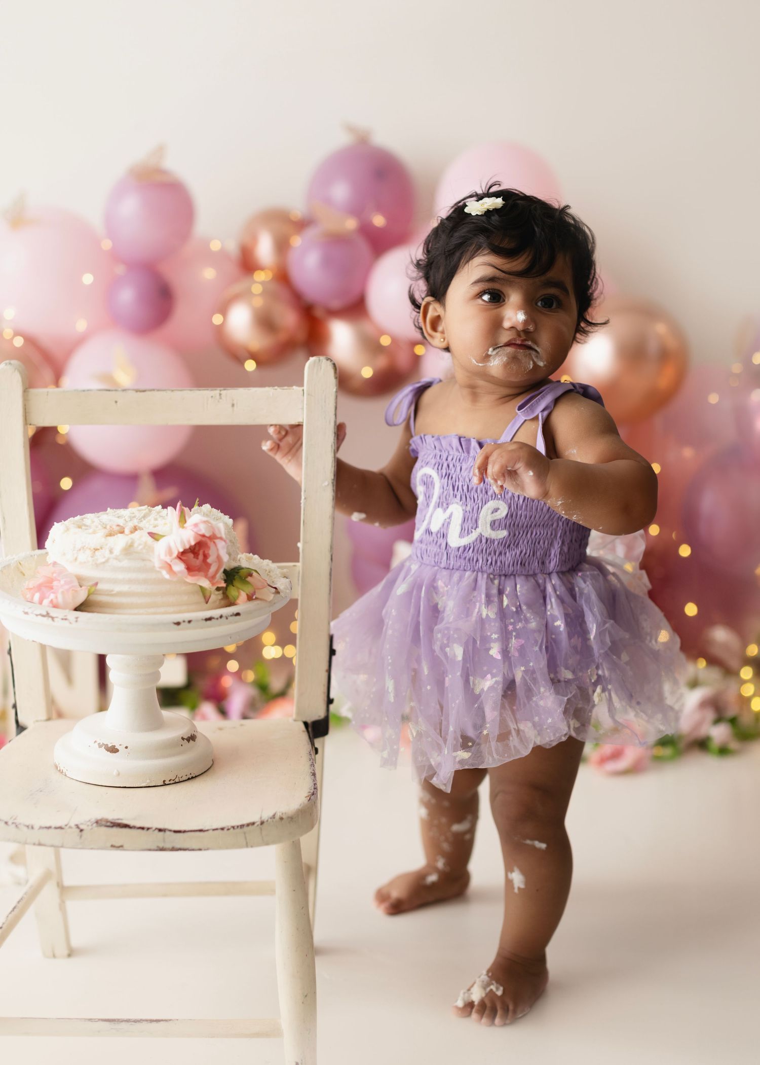 Baby girl in purple tutu dress stands by white chair with smash cake, pink balloons backdrop, first birthday photo.