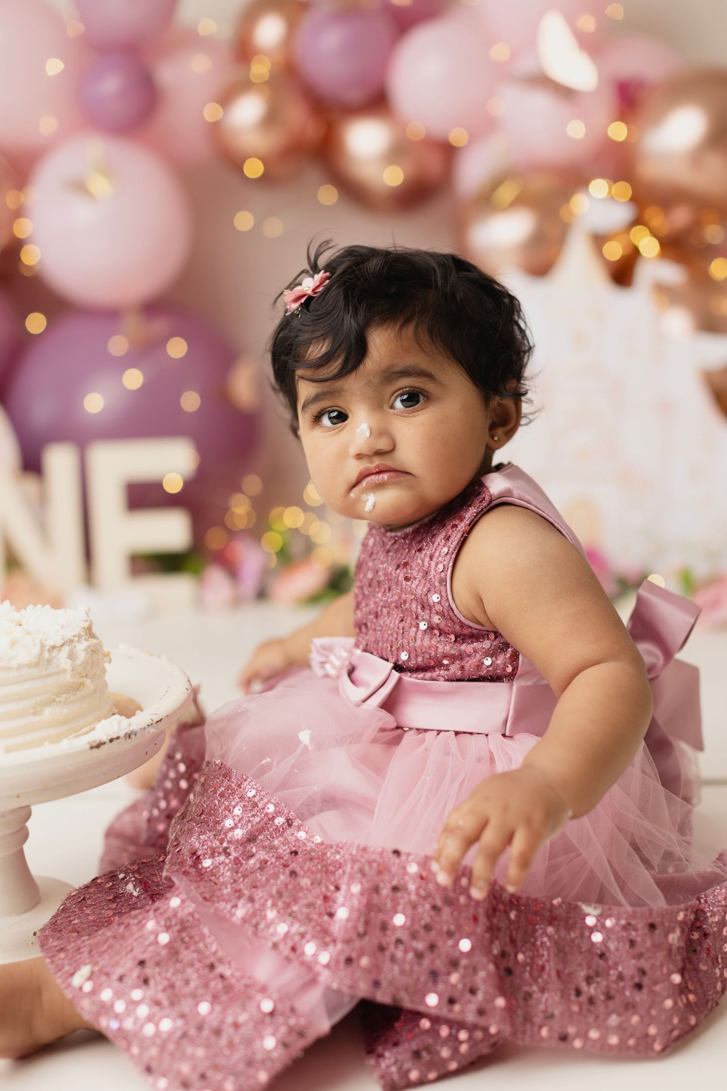 Baby girl in pink sequin dress at first birthday cake smash with balloon backdrop.