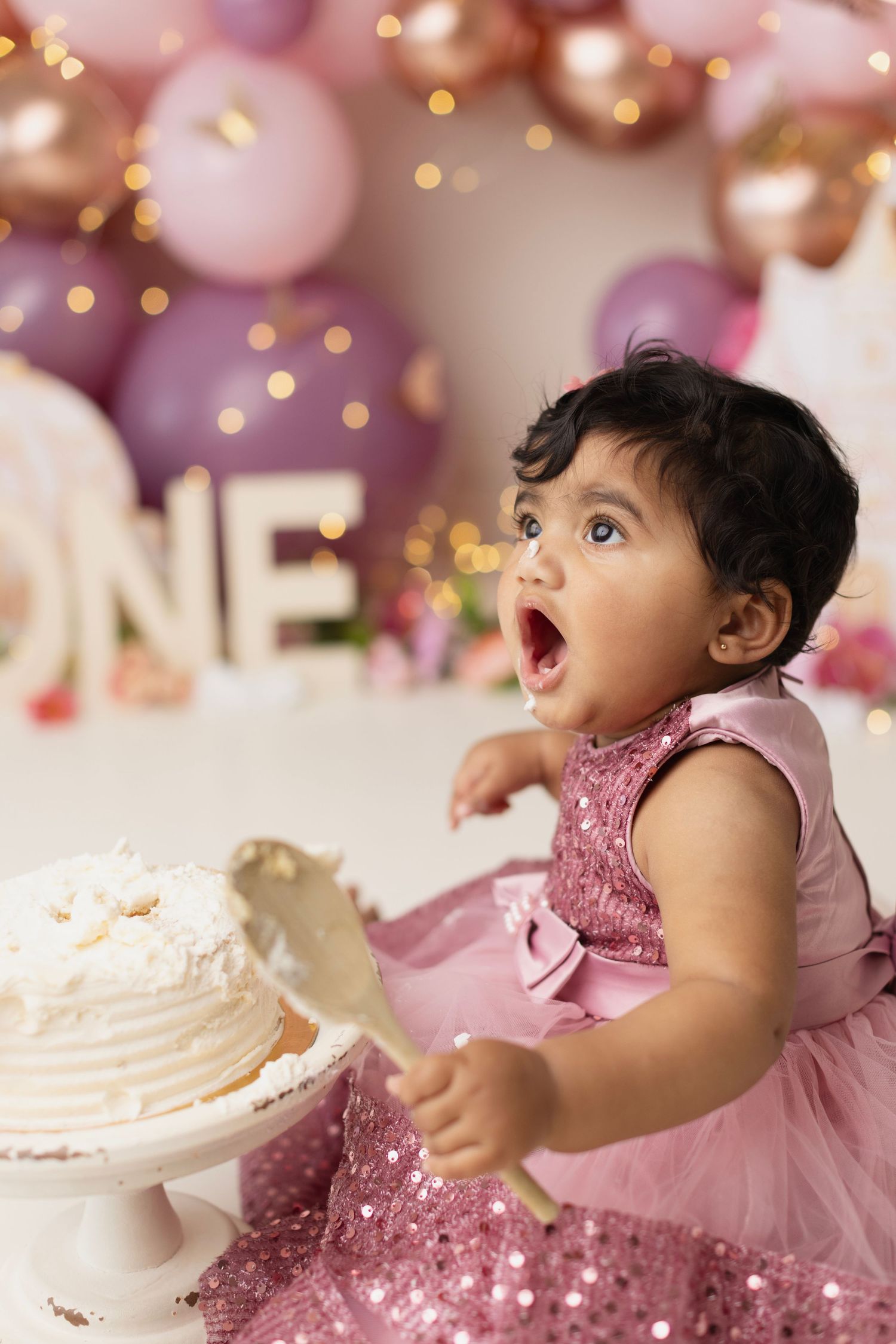Baby girl in pink sequin dress smashes first birthday cake surrounded by pink and gold balloon decorations.