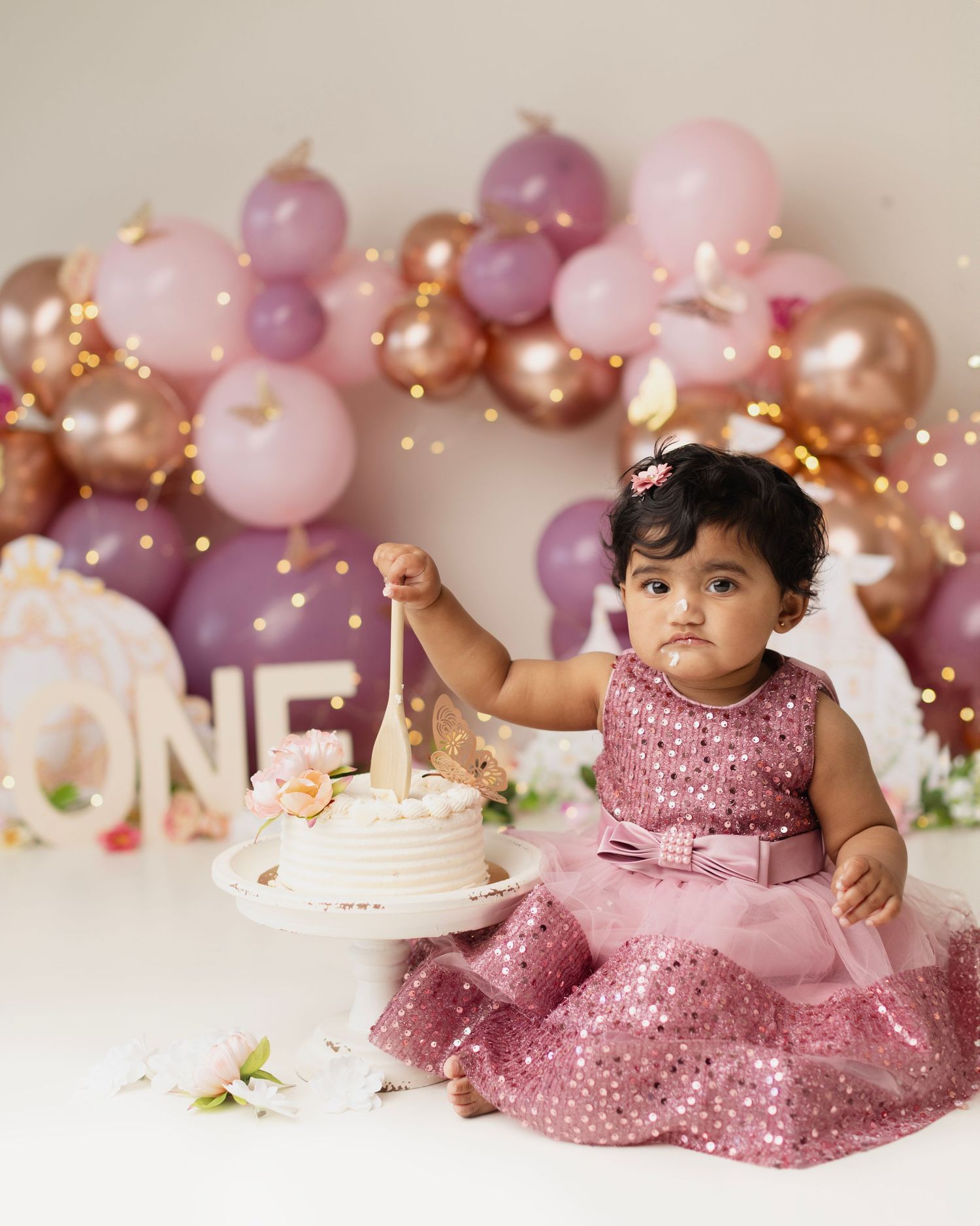 Baby girl in pink sparkle dress at first birthday cake smash with balloon garland backdrop.