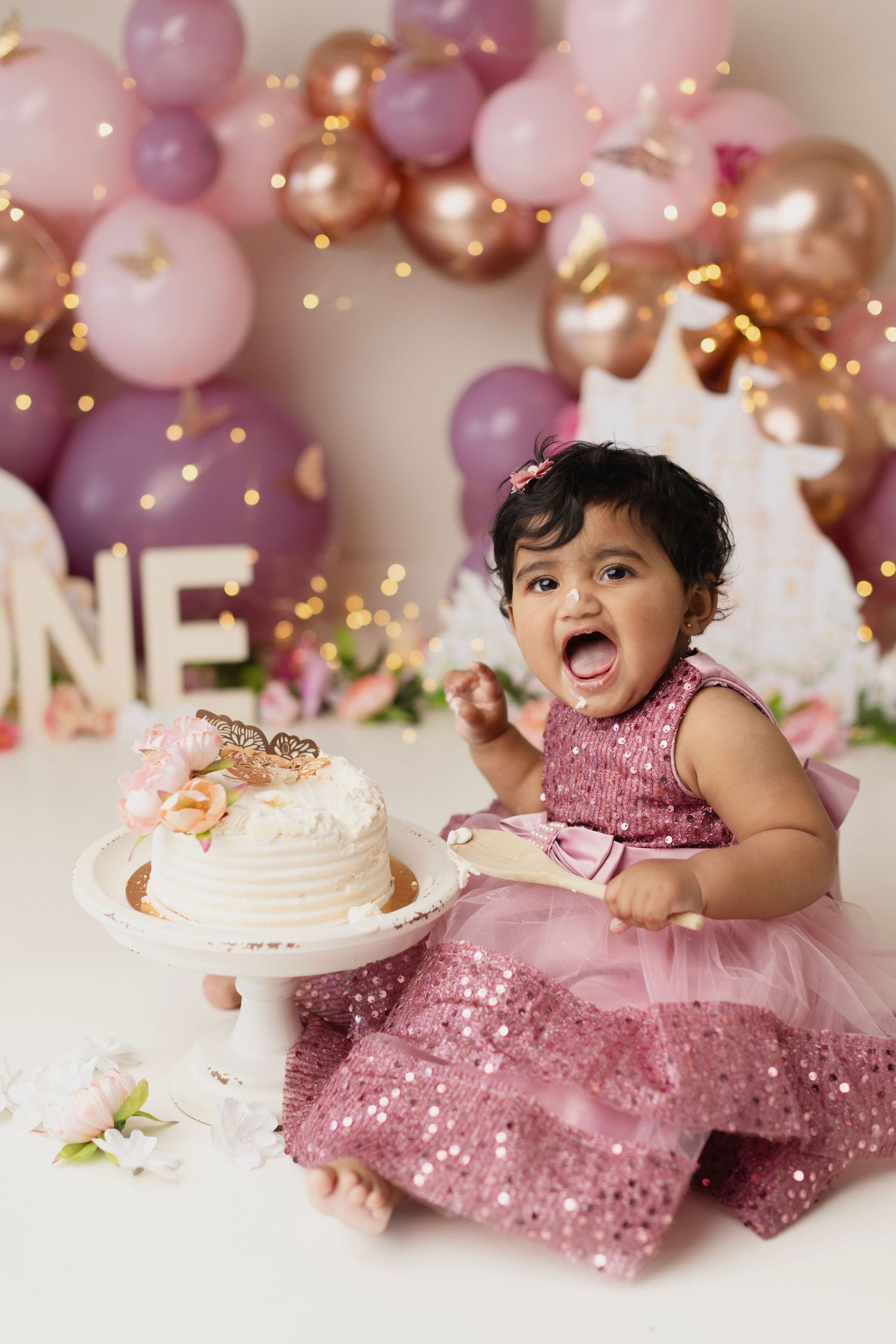 Baby girl in pink sequin dress celebrates first birthday with white cake, balloons, and ONE sign backdrop.