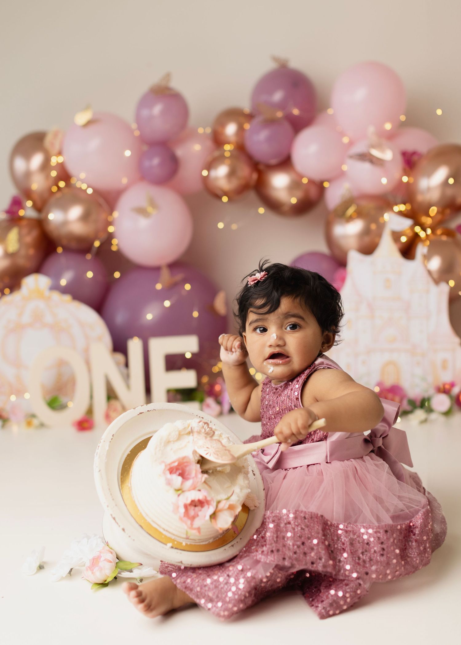 Baby girl in pink tutu dress smashes first birthday cake surrounded by pink and rose gold balloon arch.