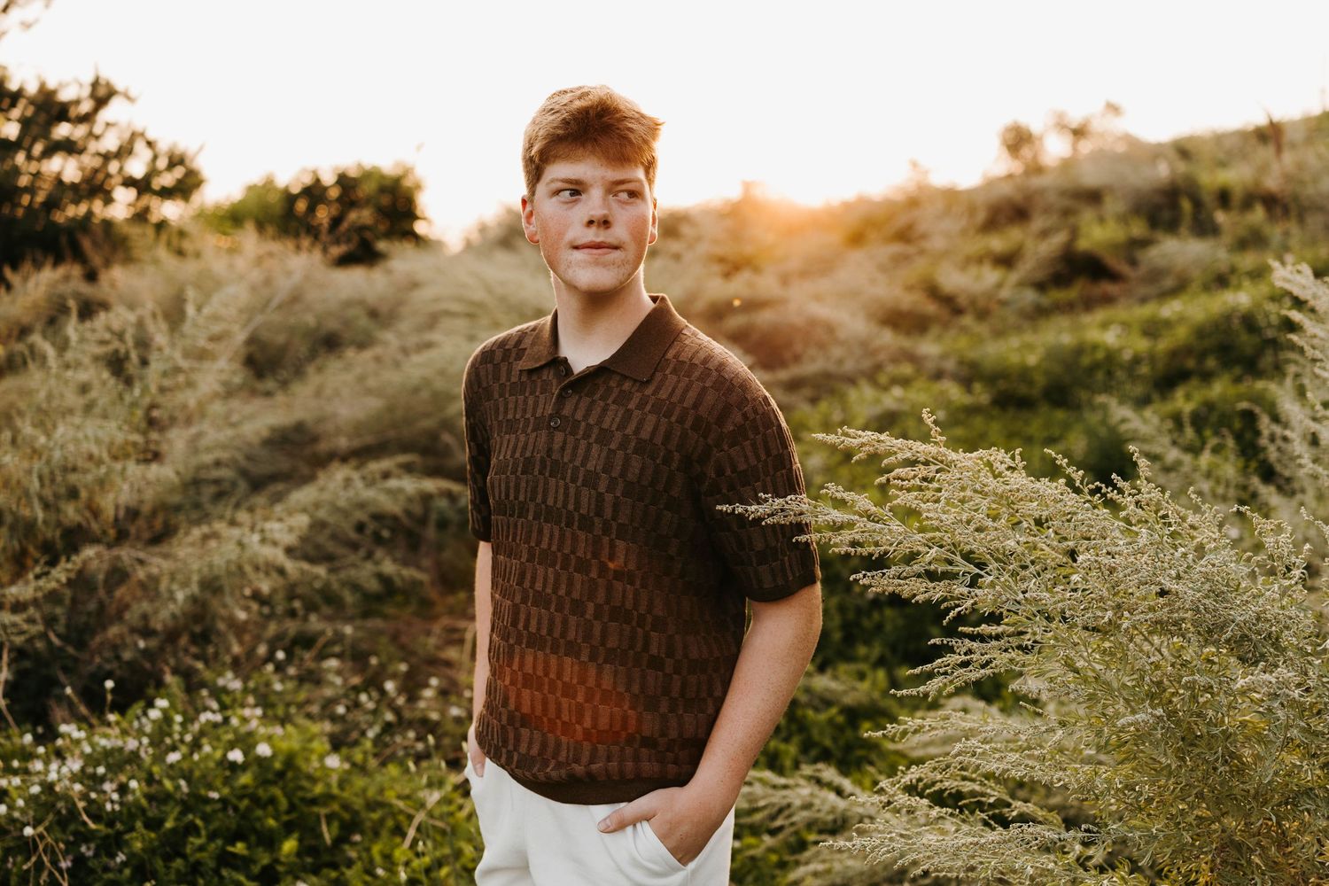 Teenage boy in brown knit polo and white pants standing in sunlit green shrubs outdoors.