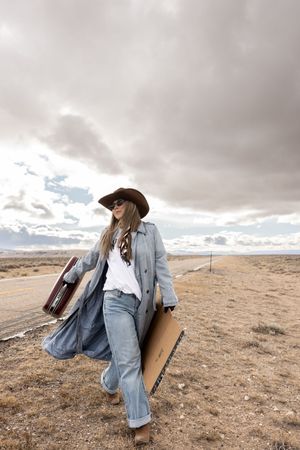 Woman in cowboy hat, trench coat, and jeans walking along a desert highway carrying luggage under cloudy skies.