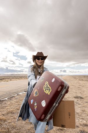 Woman in cowboy hat and sunglasses holding a sticker-covered suitcase on a desert highway under cloudy skies.