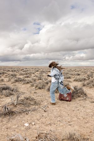 Woman in cowboy hat running through desert scrubland carrying vintage sticker-covered suitcase under dramatic cloudy sky.