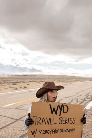 Woman in cowboy hat holding cardboard sign reading WYO Travel Series with Mercy Photography on desert road.