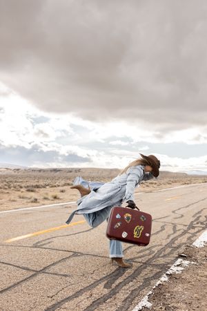 Woman in long coat balancing on desert road with vintage travel sticker suitcase under stormy clouds.