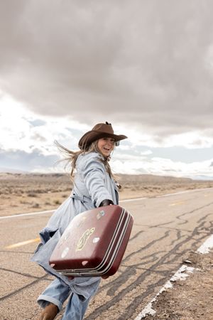 Smiling woman in cowboy hat and blue coat carrying vintage sticker-covered suitcase on empty desert highway.