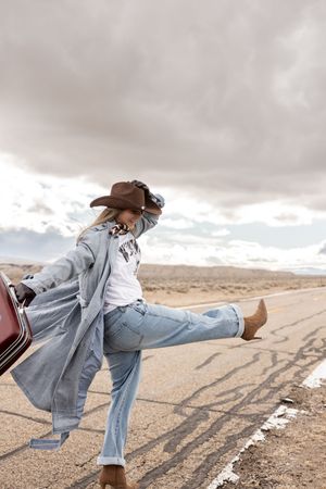 Woman in cowboy hat, duster coat, and jeans poses on desert highway beside vintage car under cloudy sky.