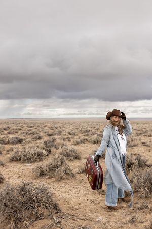 Woman in gray coat and cowboy hat carrying vintage suitcase through vast desert landscape under stormy sky.