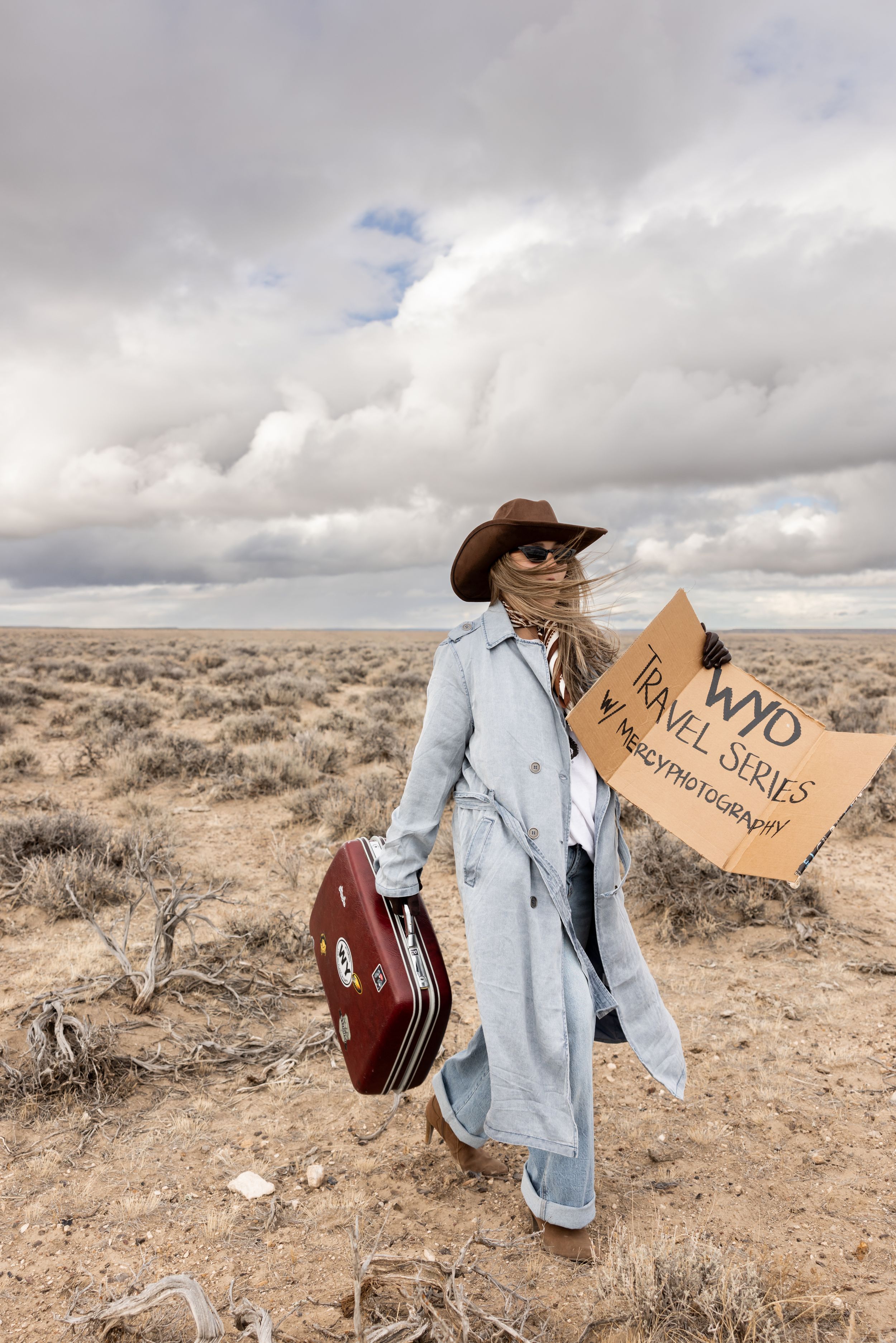 Person in trench coat and cowboy hat holds suitcase and WND Travel Series sign in vast desert landscape.