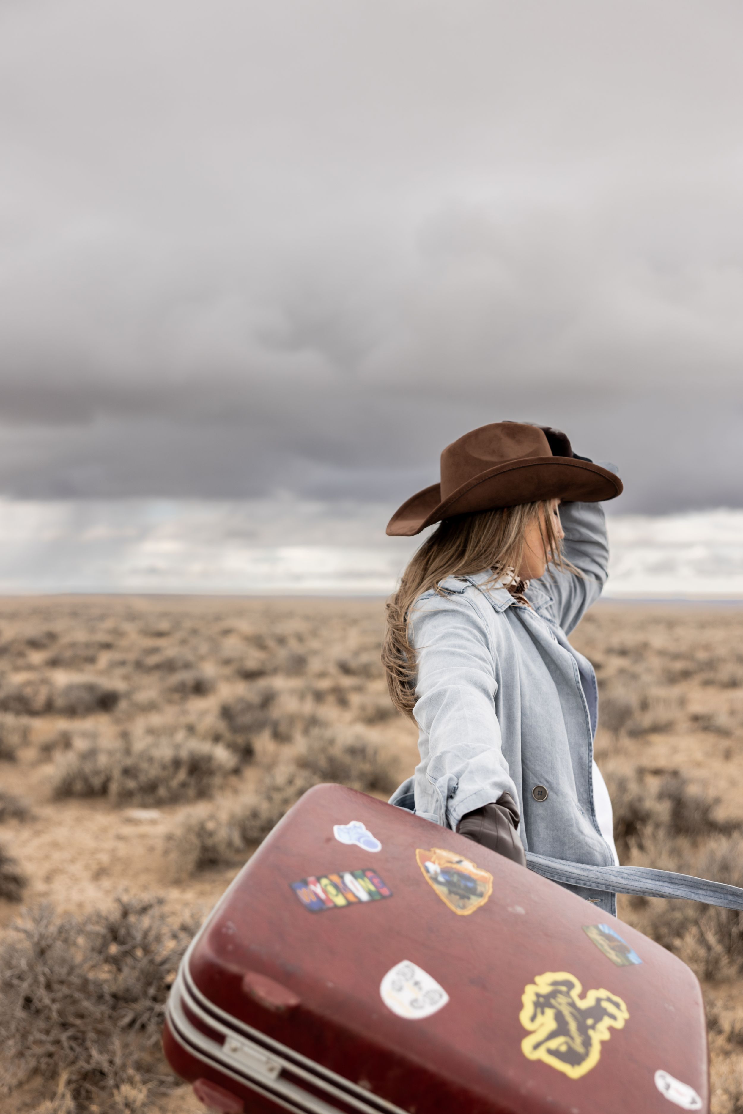 Woman in brown cowboy hat and gray coat carries sticker-covered vintage suitcase across vast desert plain.