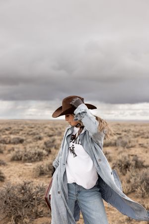 Woman in brown cowboy hat, denim jacket, and jeans poses in open desert landscape under cloudy sky.