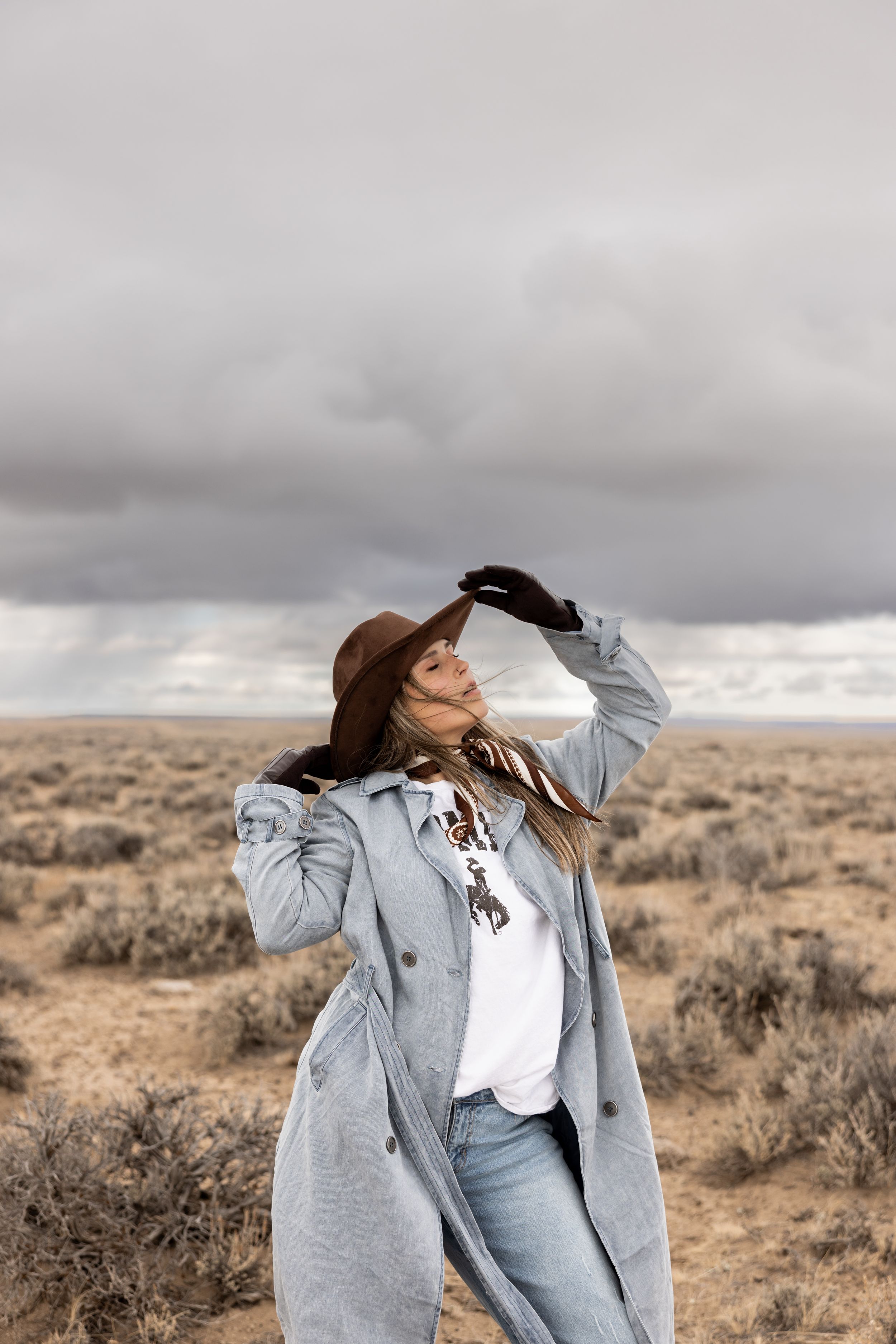 Woman in brown hat and gray coat stands in vast desert landscape under dramatic cloudy sky.