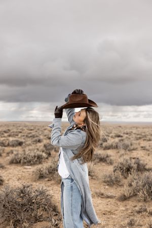 Woman in denim jacket holding brown cowboy hat in vast desert landscape under dramatic cloudy sky.