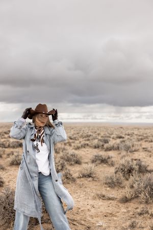 Woman in cowboy hat, denim coat, and jeans poses in arid desert landscape under dramatic cloudy sky.
