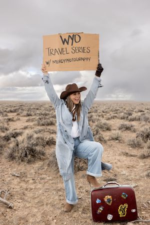 Woman in cowboy hat holds WYO Travel Series sign in desert landscape with vintage sticker-covered suitcase.