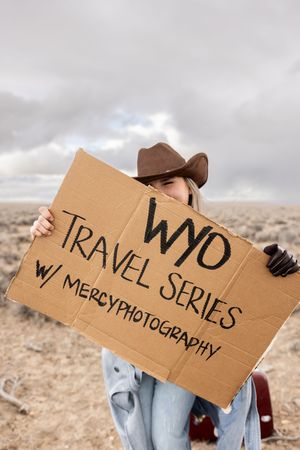 Woman in cowboy hat holds cardboard sign reading WYO Travel Series with MercyPhotography in desert landscape.