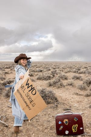 Woman in cowboy hat holding a cardboard sign in a desert landscape with a vintage sticker-covered suitcase nearby.