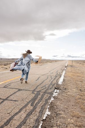 Person in cowboy hat and denim coat running along empty desert highway holding a cardboard sign.