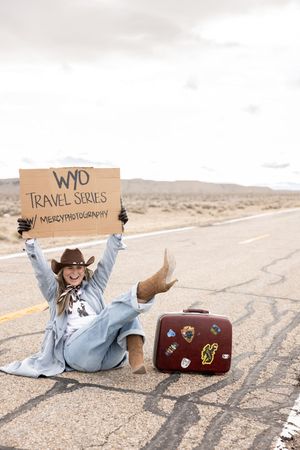Woman in cowboy hat sits on desert road holding WYD Travel Series sign beside vintage sticker-covered suitcase.