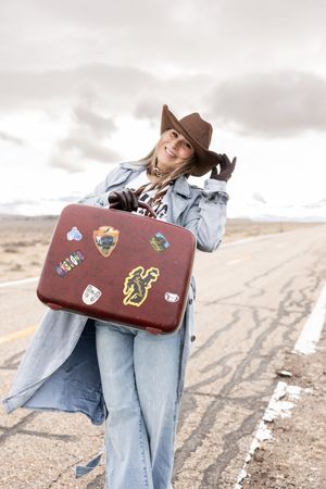 Young woman in cowboy hat and denim coat holds vintage sticker-covered suitcase on empty desert highway.