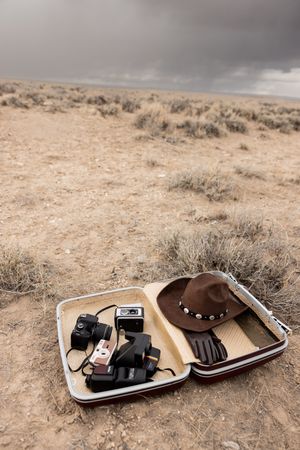 Open vintage suitcase with retro cameras, brown hat, and gloves on arid desert ground under stormy sky.