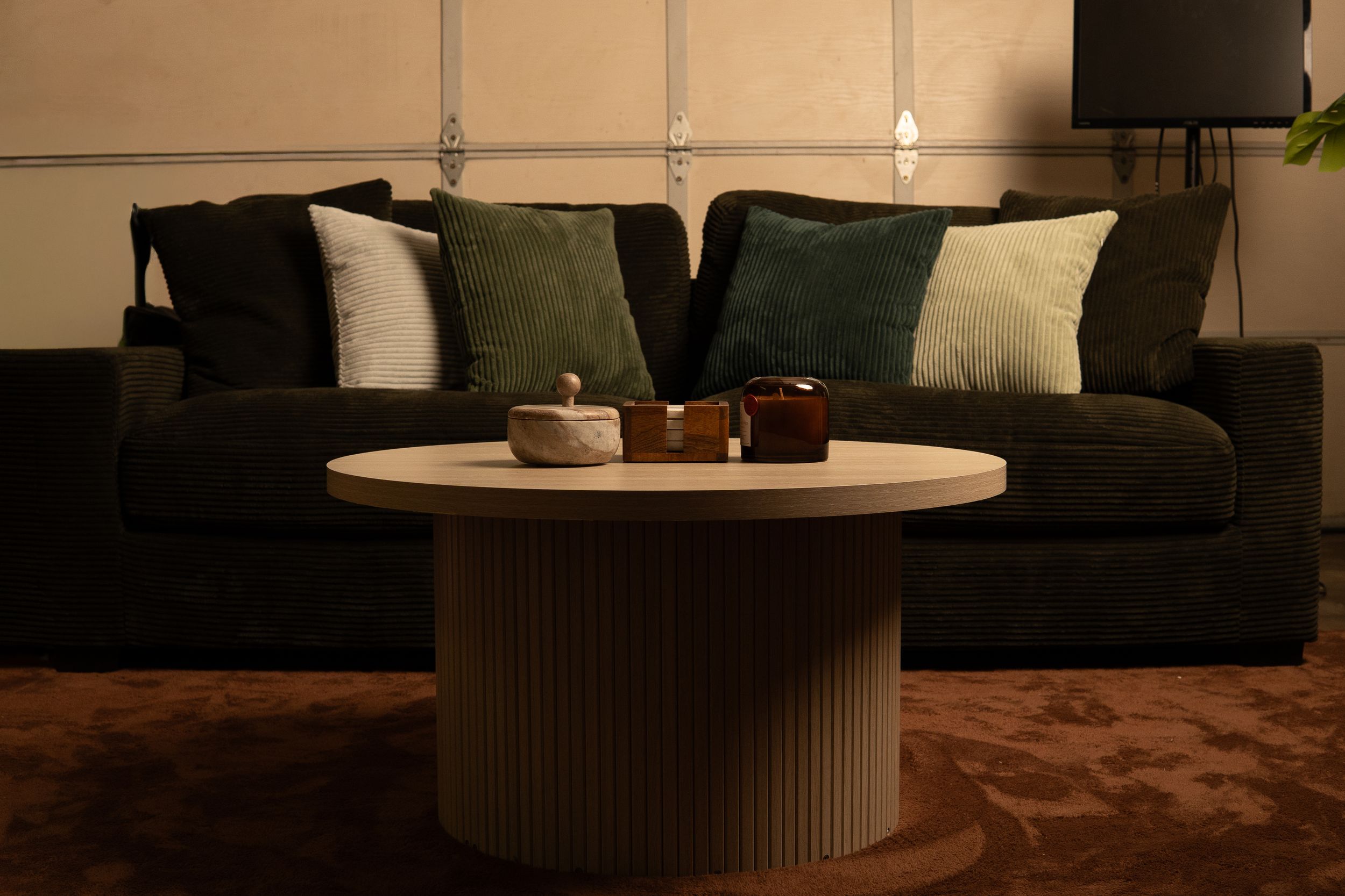 Dark green velvet sofa with green and white throw pillows behind a round ribbed coffee table with decorative items.