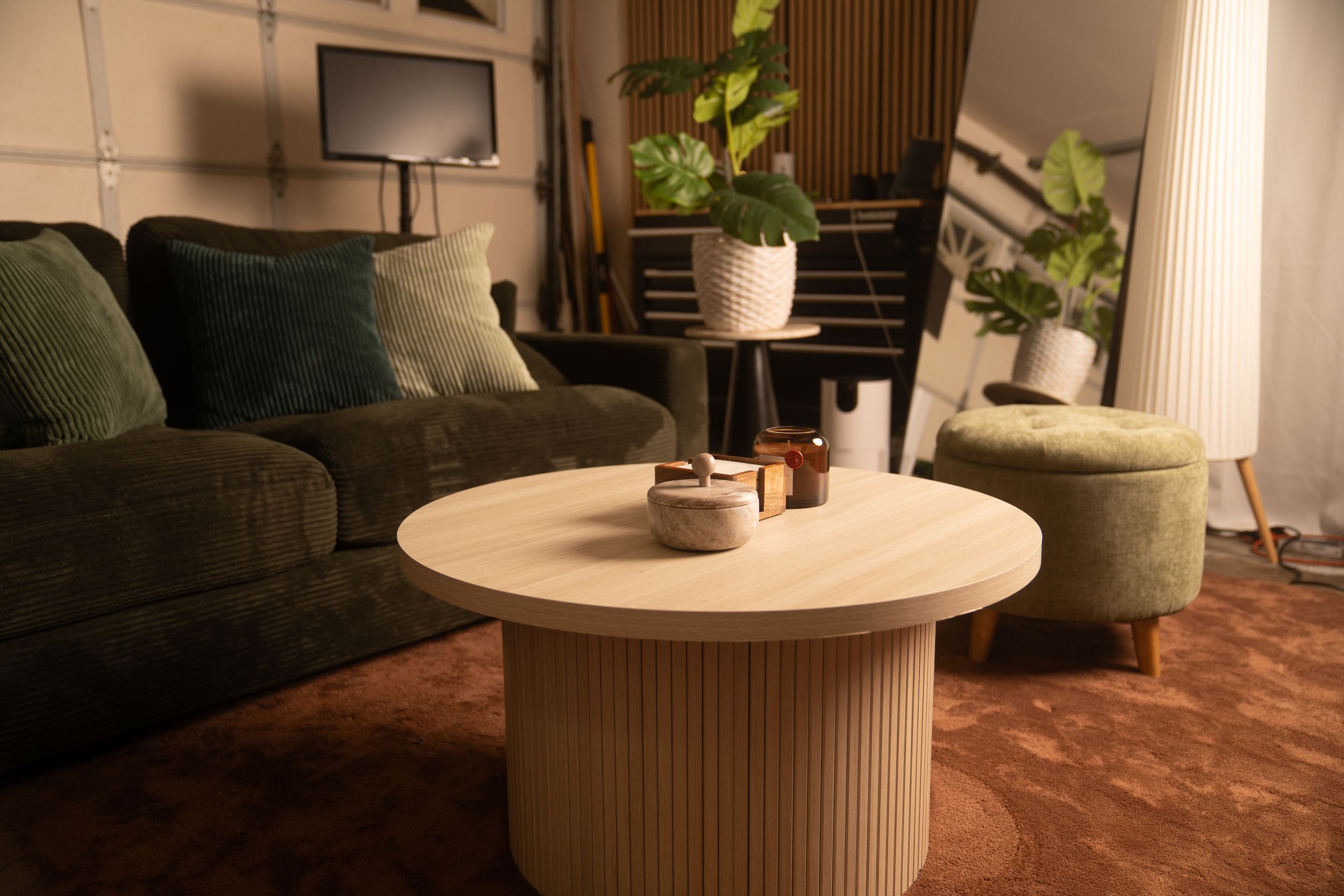 Modern living room with round fluted wood coffee table, green velvet sofa, and potted plants in warm lighting.