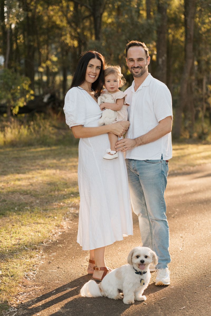 Family with baby, white fluffy dog posing outdoors on a sunny path surrounded by trees during maternity photoshoot with a dog in Rouse hill