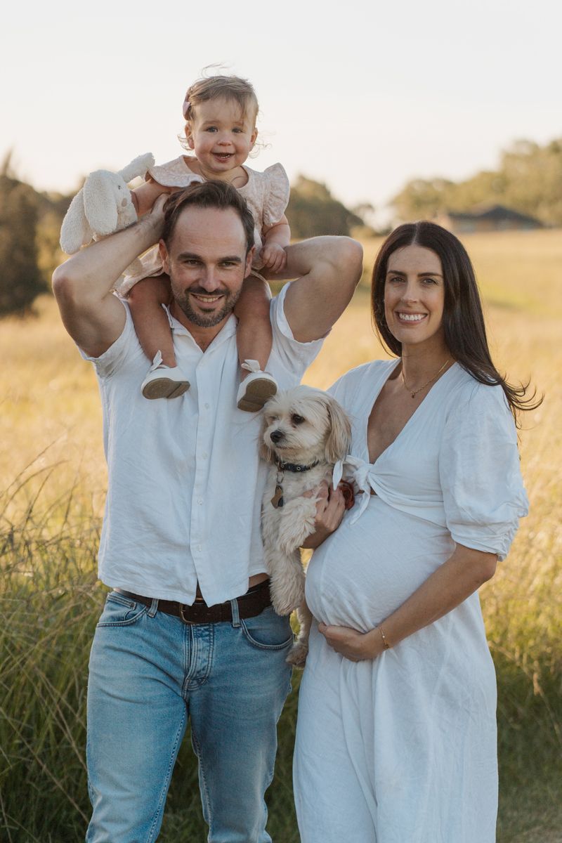 Smiling family with toddler on dad's shoulders, pregnant mom in white dress, holding a fluffy dog in a sunny field photoshoot in rouse hill